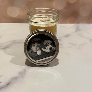 Puede incluir: Una vela en un tarro de vidrio con una foto en blanco y negro de un niño pequeño y un perro en la tapa. La foto muestra al niño y al perro mirando por una ventana.