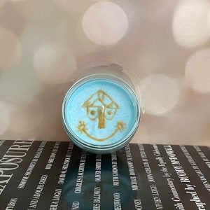 Puede incluir: Un portavelas redondo de cristal transparente con una vela azul claro en su interior. La vela tiene un diseño dorado de una casa con una cara sonriente. La vela está sobre un libro en blanco y negro.