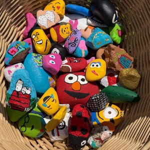 May include: A collection of small, smooth stones painted with various colourful designs, including cartoon characters, faces, and patterns, contained within a woven wicker basket. The painted rocks feature bright acrylic colours and detailed illustrations.