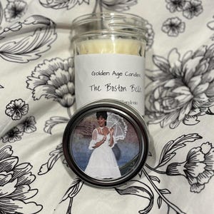 May include: A glass jar candle labeled "Golden Age Candles The Boston Belle" with a silver lid. A small, round tin with a picture of a woman in a white dress and parasol is in front of the candle. The background is a white floral pattern.