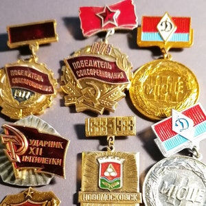 Può includere: Una collezione di medaglie e spille vintage, con varie forme e design. I toni oro, rosso e argento sono prominenti. Alcuni pezzi includono testo e simboli, come stelle, bandiere e la falce e il martello.
