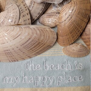 May include: A collection of seashells in various shades of brown and beige, arranged on a striped blue and yellow fabric with the text "the beach is my happy place" embroidered on it.