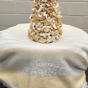 May include: A decorative tree sculpture made of various seashells in shades of beige and cream. The tree sits on a striped towel with the embroidered text "the beach is my happy place".