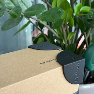 May include: A rectangular cardboard box featuring two black corner protectors with the embossed text RDPrint. The top surface of the box displays the printed word Accessory. The box is constructed from light brown paperboard material.