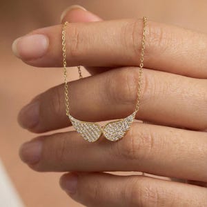 Op de afbeelding: Een gouden ketting met een hanger in de vorm van engelen vleugels. De vleugels zijn versierd met kleine, sprankelende kristallen. De ketting wordt in een hand gehouden, waarbij de fijne ketting en het ontwerp van de hanger worden benadrukt.