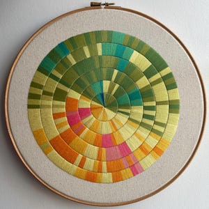 May include: A circular embroidery hoop with a colorful geometric design. The design is made up of concentric circles of different colors, including green, yellow, orange, pink, and blue.