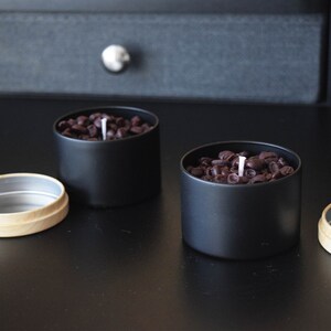 May include: Two black candle jars with a white wick. The jars are filled with brown coffee beans.