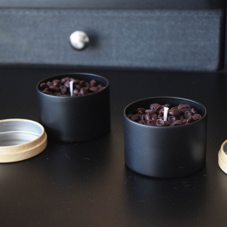 May include: Two black candle jars with a white wick. The jars are filled with brown coffee beans.
