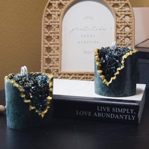May include: Two green and gold candles with a crystal-like texture. The candles are sitting on a black book with the text "Live Simply, Love Abundantly".