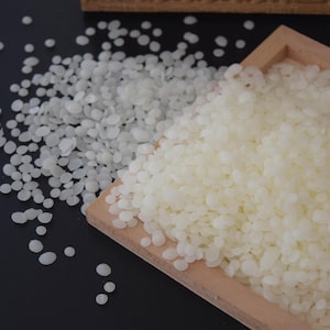May include: White wax beads in a wooden tray. The beads are small and round, and they are scattered on a black surface.