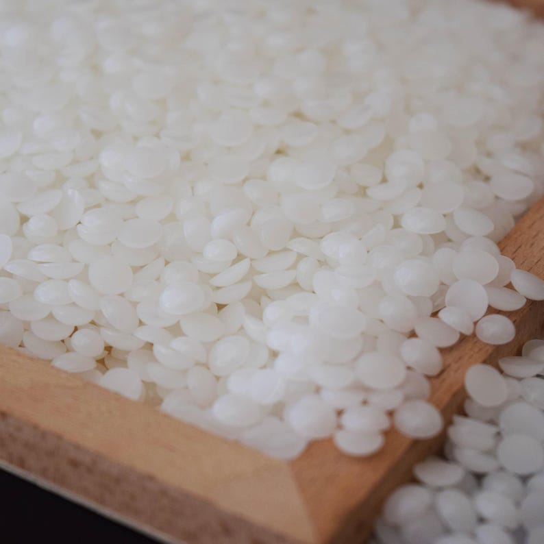 May include: Close-up of a pile of small, off-white, disc-shaped wax beads. The beads are contained within a light brown wooden tray. The wax beads have a smooth, slightly glossy appearance, suggesting they are a cosmetic or crafting material.