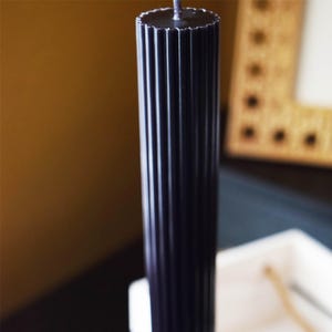 May include: A tall, black pillar candle with vertical ribbed detailing. The candle has a scalloped top and a wick extending from the center. The candle is a decorative item for home decor.