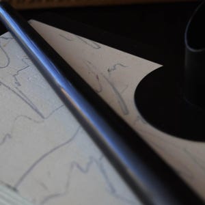 May include: A black, cylindrical object, possibly a pen or a candle holder, lies diagonally across a textured, off-white surface with faint blue markings. A black, circular object is visible in the background, suggesting a minimalist design.