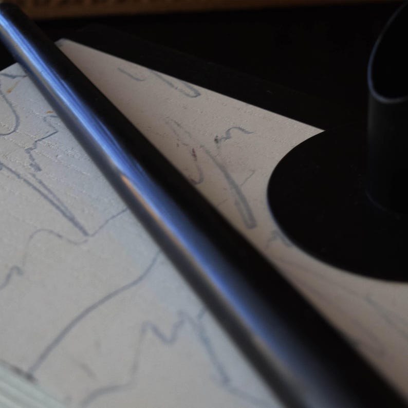 May include: A black, cylindrical object, possibly a pen or a candle holder, lies diagonally across a textured, off-white surface with faint blue markings. A black, circular object is visible in the background, suggesting a minimalist design.