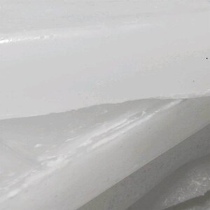 May include: Close-up of a white, textured surface with a layered appearance. The image shows a diagonal cut or break, revealing a slightly different texture underneath. The lighting creates highlights and shadows, emphasizing the surface's details.
