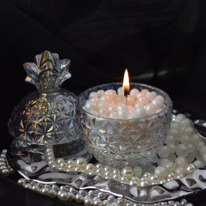May include: A lit candle in a clear, patterned glass bowl filled with white beads. A decorative pineapple-shaped glass container sits nearby. The items are on a silver tray with pearl strands, set against a dark background.