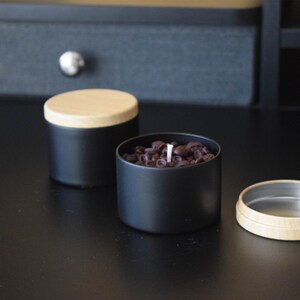 May include: Two black candle jars with wooden lids. One jar is filled with coffee beans and has a white candle wick. The other jar is empty.