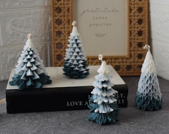 May include: Four Christmas tree-shaped candles in varying sizes and shades of white and teal. The candles are arranged on a black book with the words "LOVE ALWAYS" visible. A decorative frame is in the background.