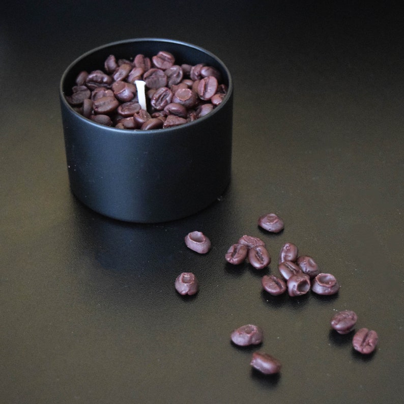 May include: A black candle jar filled with coffee beans and a white wick. The jar is sitting on a black surface with several coffee beans scattered around it.