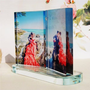 May include: A glass photo frame with two separate panels, each displaying a photo of a couple on a beach. The frame is on a clear glass base. The text "I now" is visible on the right panel.