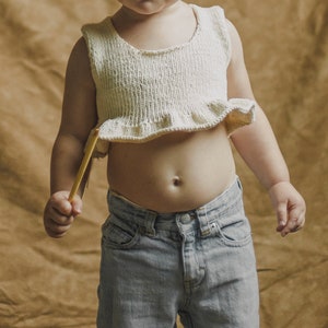 May include: A young child wearing a white crocheted top with a ruffled hem and light blue denim jeans.