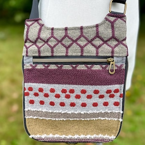 May include: A crossbody bag featuring a textured fabric with a geometric pattern in maroon and gray at the top. The lower section displays horizontal bands of maroon, gray with red dots, and a solid gold panel, all secured with a black zipper.