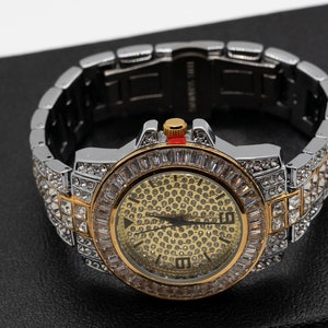 Könnte beinhalten: Eine silber- und goldfarbene Armbanduhr mit rundem Zifferblatt, besetzt mit klaren Kristallen. Die Uhr hat ein goldfarbenes Zifferblatt mit kleinen, dunklen Punkten und ein silbernes Armband. Die Uhr wird auf einer schwarzen Oberfläche präsentiert.
