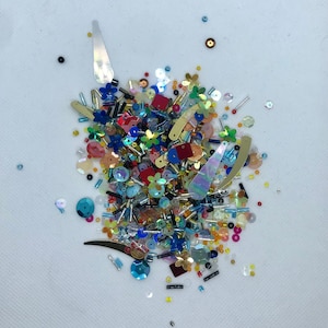 May include: A vibrant collection of craft embellishments, including sequins, beads, and decorative shapes. The mix features a variety of colours, such as red, blue, gold, and iridescent elements. Shapes include flowers, rectangles, and teardrops, ideal for craft projects.
