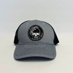 May include: A gray trucker hat with a black mesh back. The front features a circular patch with a skull, flowers, and the words "MEMENTO MORI" in a black and white design. The hat has a curved brim.