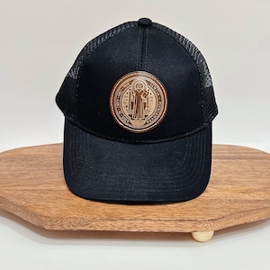 May include: Black trucker hat with a mesh back and a curved brim. The front features a circular, brown leather patch with a religious emblem. The hat is displayed on a wooden, oval-shaped tray.