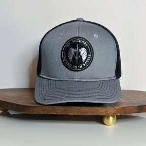 May include: A gray and black trucker hat with a circular patch featuring the text "ST. MICHAEL" and "DEFEND US IN BATTLE". The patch includes a design of wings and a sword. The hat is displayed on a wooden tray.