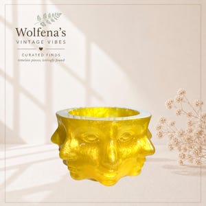May include: A gold-colored, vintage-style decorative bowl with three faces. The bowl has a white rim and the text "Wolfena's Vintage Vibes" is visible in the background. The bowl is a unique piece for home decor.