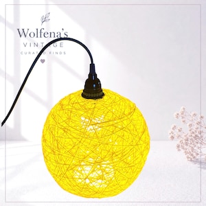 May include: A spherical pendant lamp shade crafted from bright yellow intertwined string or twine. The lamp features a black socket and a black electrical cord extending from the top. The text Wolfena's VINTAGE CURATED FINDS is visible.