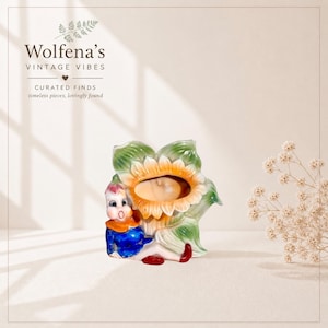 May include: A vintage ceramic figurine featuring a sunflower with a baby in the center. A small figure in blue, orange, and red clothing sits next to the flower. The text "Wolfena's Vintage Vibes" is at the top.