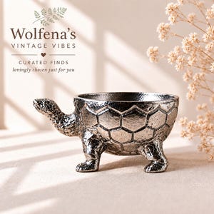 May include: A metallic silver turtle-shaped bowl featuring a textured shell with a hexagonal pattern, a detailed head, and four sturdy legs. The bowl has a smooth interior and a reflective finish. Text above reads Wolfena's VINTAGE VIBES.