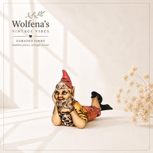 May include: A vintage ceramic gnome figurine, lying down with hands supporting the head. The gnome wears a red hat, red and yellow shirt, and black shoes. The text "Wolfena's Vintage Vibes" is in the upper left corner.