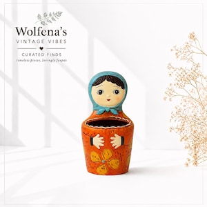 May include: A ceramic wall pocket shaped like a nesting doll with a blue headscarf, dark hair, and a painted face. The body is orange with yellow floral patterns and white hands. Text above reads Wolfena's VINTAGE VIBES CURATED FINDS.