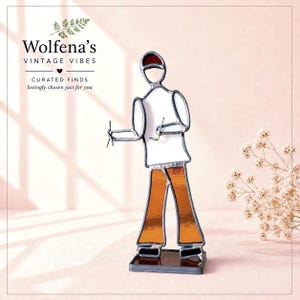 May include: Stained glass figure of a person in a white shirt and orange pants, holding tools. The figure has a red hat and is standing on a rectangular base. The figure is a vintage item.