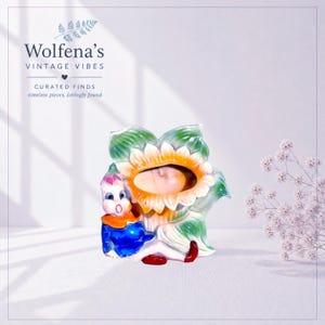 May include: A ceramic figurine featuring a small character with a pink-tinted head and blue shirt sitting beside a large, yellow-petaled sunflower with green leaves. The sunflower center forms an oval opening. Text reads Wolfena's VINTAGE VIBES.