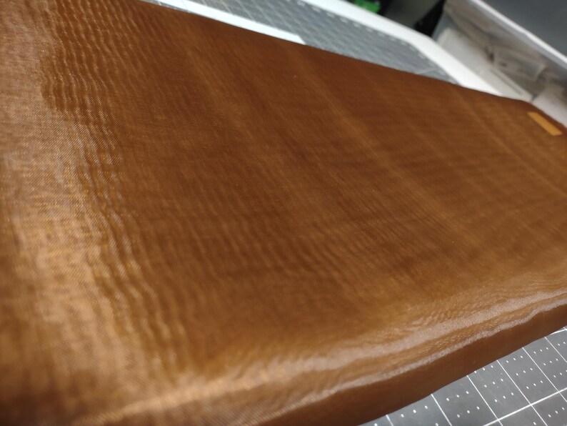 May include: A brown, textured, rectangular fabric with a subtle sheen. The fabric is smooth and appears to be made of a fine mesh.