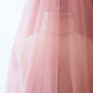 May include: A pale pink tulle fabric with a soft, delicate texture. The fabric is sheer and lightweight, with a subtle sheen.