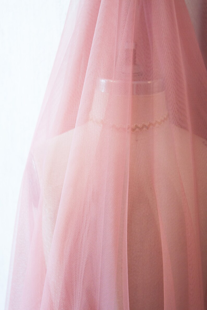 May include: A pale pink tulle fabric with a soft, delicate texture. The fabric is sheer and lightweight, with a subtle sheen.