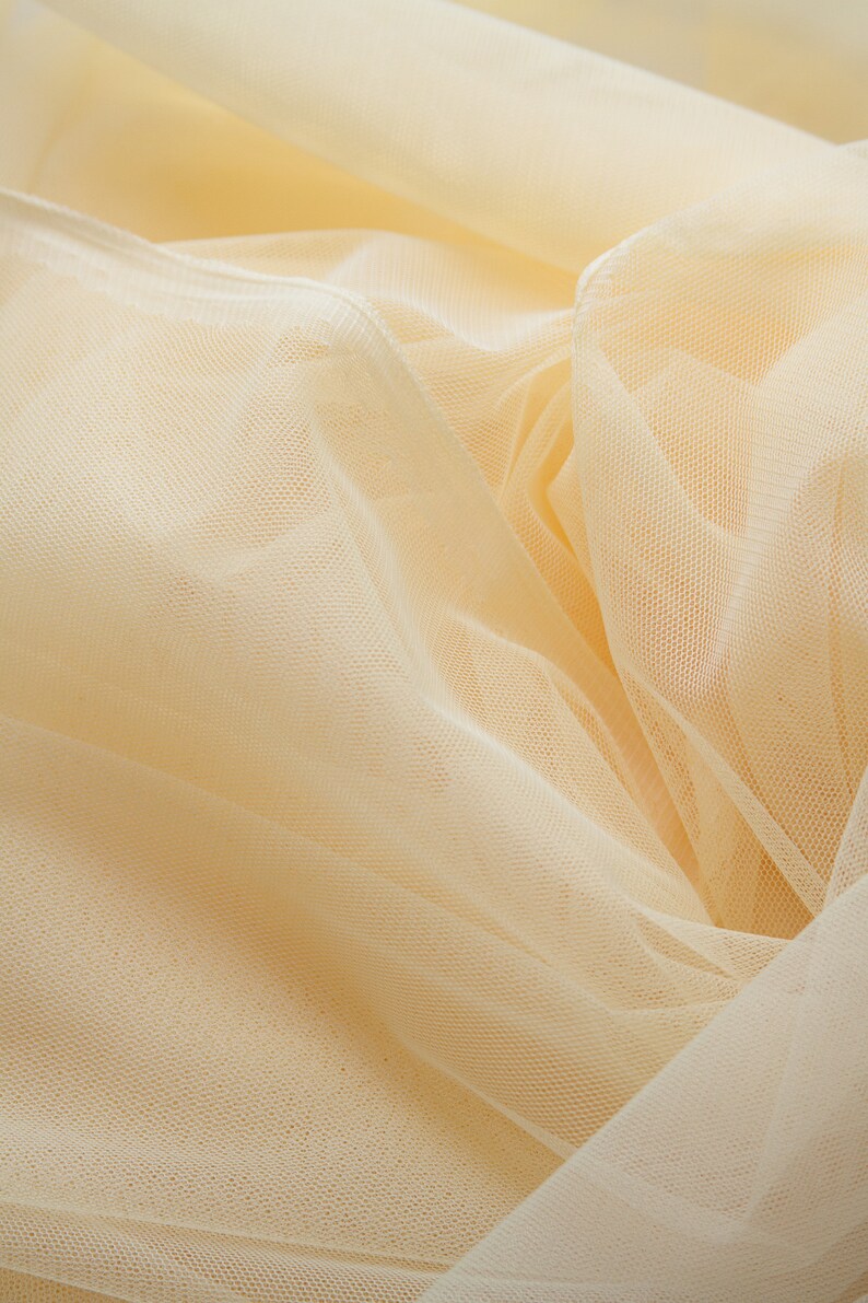May include: A close-up of a soft, pale yellow tulle fabric. The fabric is delicate and has a fine mesh texture.
