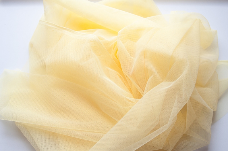 May include: A pale yellow tulle fabric, soft and delicate, with a slightly crinkled texture. The fabric is draped and folded, creating a flowing and airy appearance.