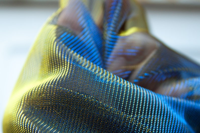 May include: Close-up of a blue and gold fabric with a textured, iridescent finish. The fabric has a subtle pattern of small dots.