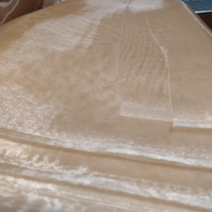 May include: A close-up of a sheer, light brown fabric with a subtle shimmer. The fabric is folded in layers, creating a soft, flowing texture.