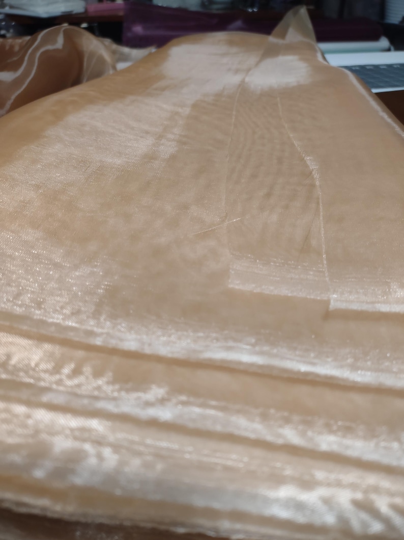 May include: A close-up of a sheer, light brown fabric with a subtle shimmer. The fabric is folded in layers, creating a soft, flowing texture.