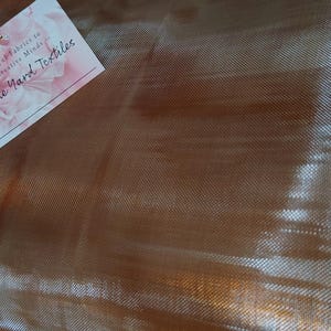 以下が含まれることがあります： 細かい織り目のメッシュテクスチャを持つ、メタリックな銅色の生地のロール。生地の上には、花のグラフィック、「Beautiful Fabrics for Creative Minds」というテキスト、およびブランド名「By The Yard Textiles」が記載された白い長方形のタグが置かれています。