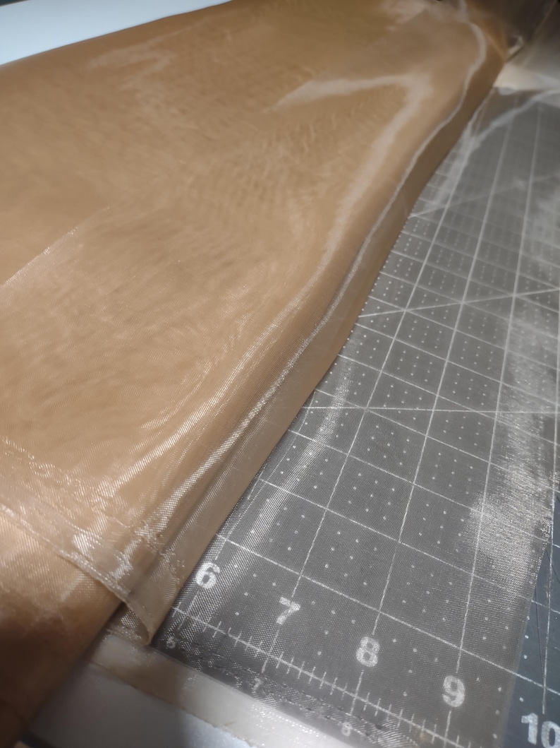 May include: A roll of beige sheer fabric with a slight sheen. The fabric is folded in half and the edge is visible. The fabric is lying on a cutting mat with a grid pattern.