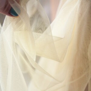 May include: Close-up of a sheer, ivory-colored tulle fabric. The fabric is soft and delicate, with a slightly textured surface.
